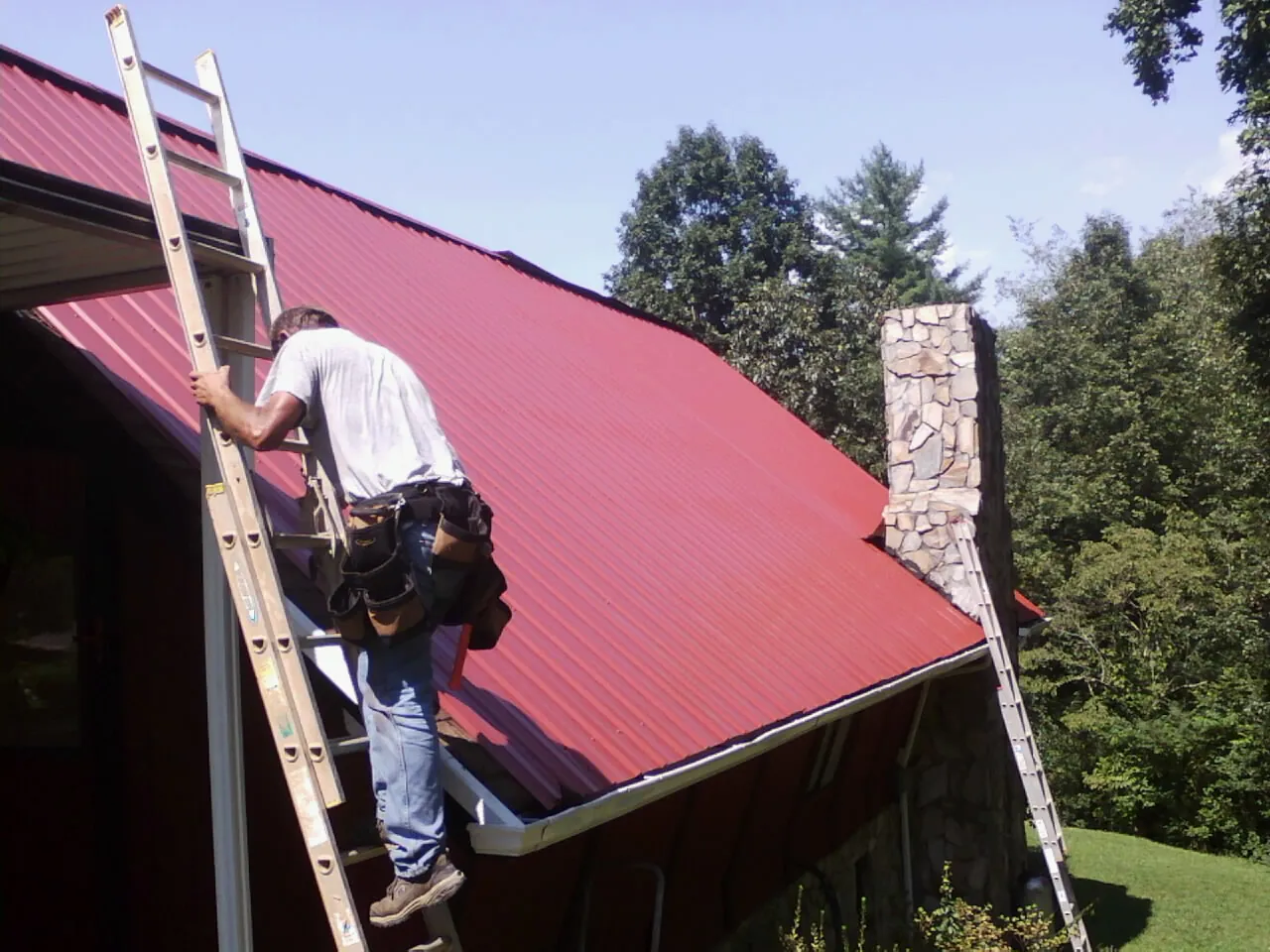 Roofer on a ladder beside a red metal roof and stone chimney — Soffit & Fascia Repair by Roofing Brothers in Ochlocknee